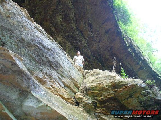 Man stands on a rocky ledge beneath a large rock overhang with greenery above