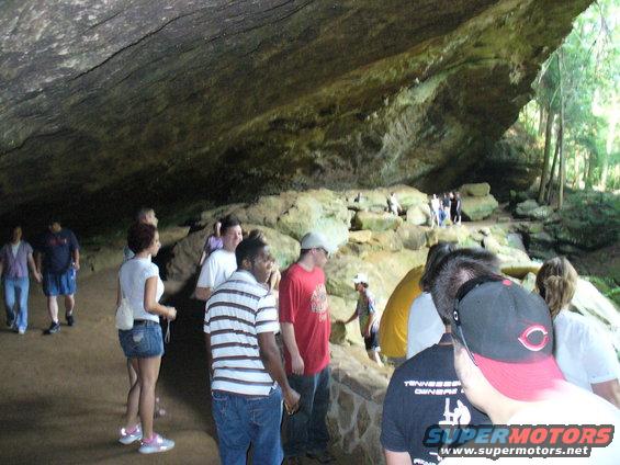 People explore a large cave with a rocky ceiling and stone walls, some wearing casual clothes