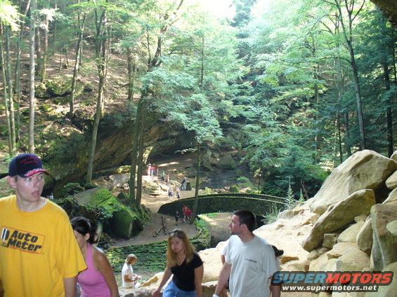 People walk near a stone bridge in a forest with large rock formations and trees
