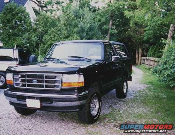 Black Ford Bronco parked on a gravel driveway, front 3/4 view. Another vehicle is partially visible to the left