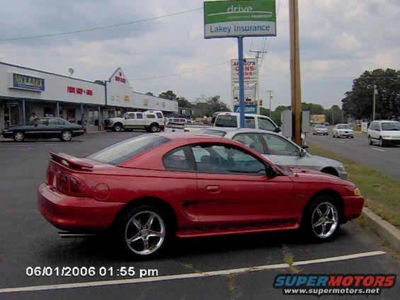 Red 1996 Ford Mustang parked next to a silver sedan, with a strip mall and signs in the background