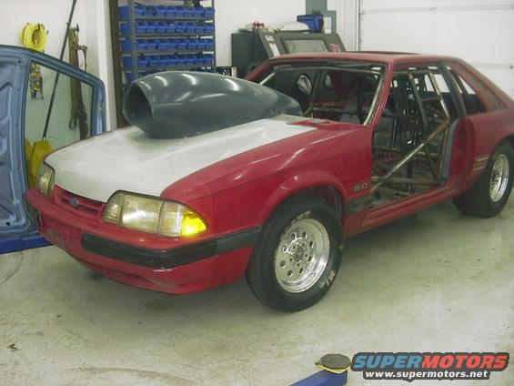 Red Ford Mustang with a large hood scoop and roll cage, missing doors and interior