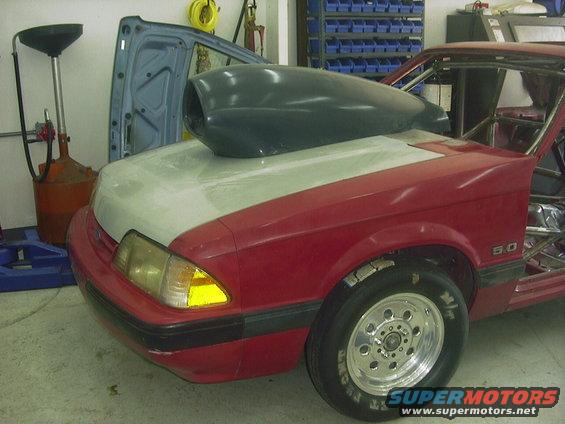Red race car, looks like a 1990 Ford Mustang, with a large hood scoop and roll cage. Front fender has a '5.0' badge