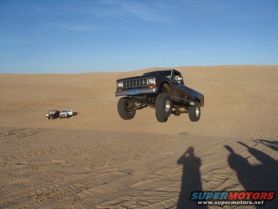 Brown pickup truck mid-air jump over sand dune, with two other trucks in the background