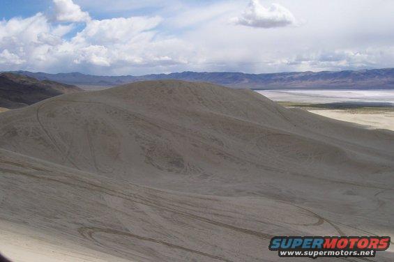 Wide shot of sand dunes with tire tracks, mountains in background, and a dry lake bed