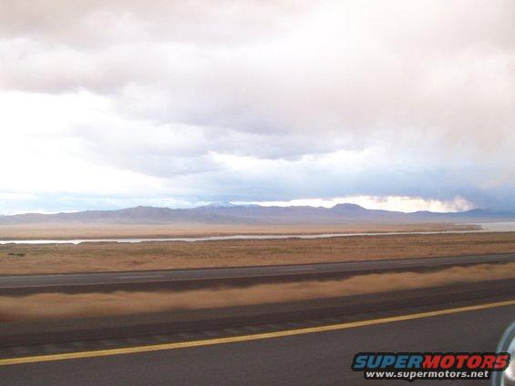 Highway passes through a desert landscape with distant mountains under a cloudy sky. A sliver of a vehicle is visible on the right
