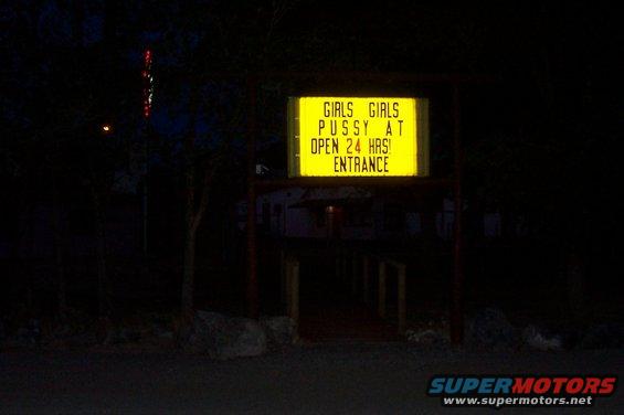 Yellow illuminated sign at night reads 'GIRLS GIRLS PUSSY AT OPEN 24 HRS! ENTRANCE'