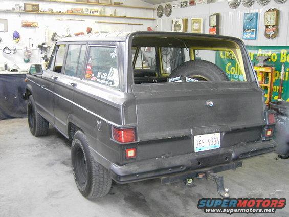 Rear view of a dark-colored Jeep Grand Wagoneer parked in a garage. It has a spare tire mounted inside the rear cargo area and a trailer hitch