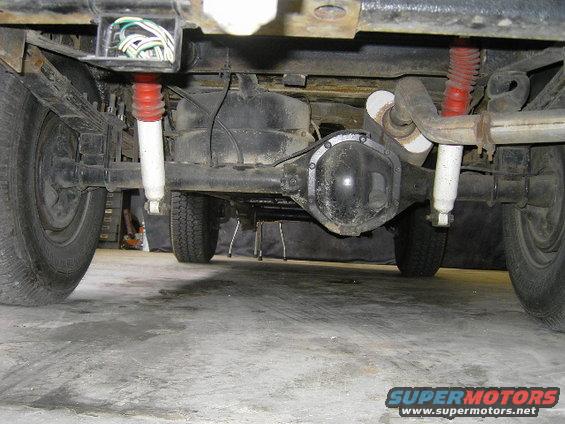 Underside view of a vehicle's rear axle, leaf springs, and shocks. Shows a fuel tank and exhaust system