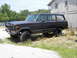 1977 Jeep Wagoneer - A dark gray 1977 Jeep Wagoneer is parked on a gravel lot next to a building.