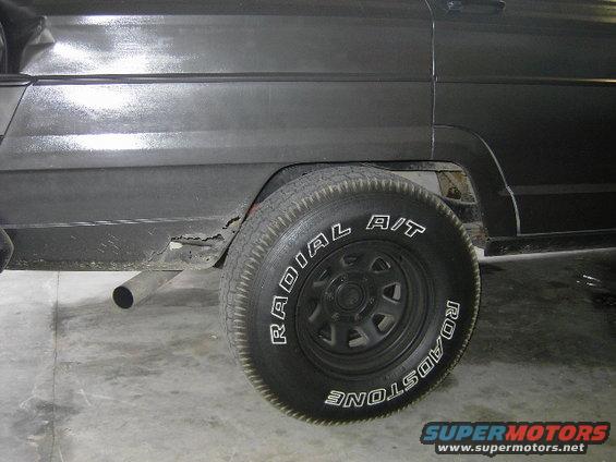 Close-up of a dark-colored Jeep Wagoneer's rear wheel well, showing rust on the lower body panel and a Roadstone Radial A/T tire