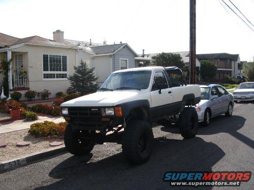 Lifted white pickup truck with large tires parked on street, silver sedan behind it