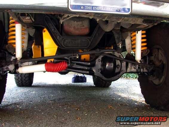 Undercarriage view showing suspension components, steering linkage, and rear axle. A blue bag is on the ground