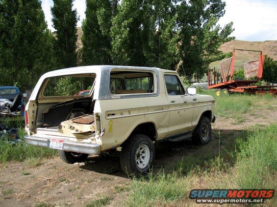 Cream-colored Ford Bronco with a white roof, viewed from the rear-left. The tailgate is open, revealing the interior. The vehicle shows signs of wear and tear