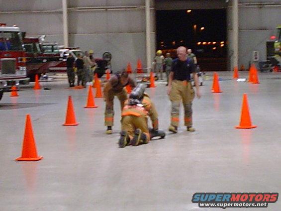 Firefighters in gear practice around orange cones inside a large building, with fire trucks visible in the background