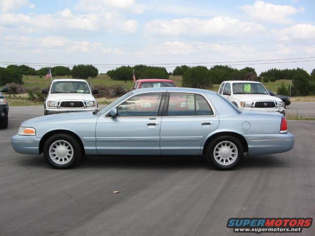 Light blue 2002 Ford Crown Victoria sedan, side view. Two white trucks and a red vehicle in background