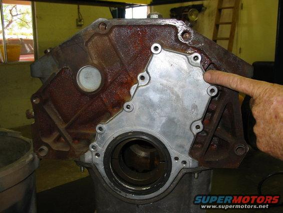 Close-up of a rusty engine block with a metal plate attached, finger pointing to a bolt hole