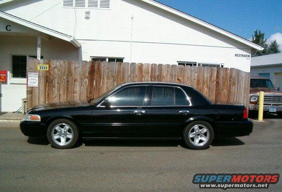 Black 2002 Ford Crown Victoria sedan parked on asphalt, side view. A brown pickup truck is visible behind a wooden fence
