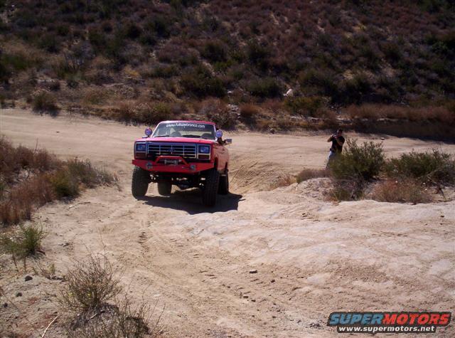 Red 1980s Ford Bronco modified for off-roading, driving on a dirt trail with a person in the background