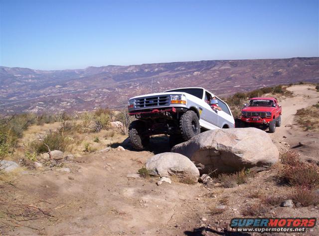 White 1995 Ford Bronco climbing over rocks on a mountain trail with a red Ford pickup behind it