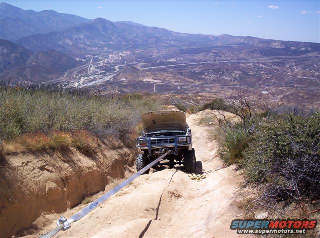 Blue Ford Bronco with hood open, stuck on a dirt trail, attached to a tow strap