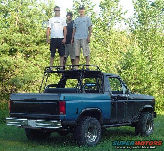 Ford Bronco with custom roll cage and roof rack, three men standing on top