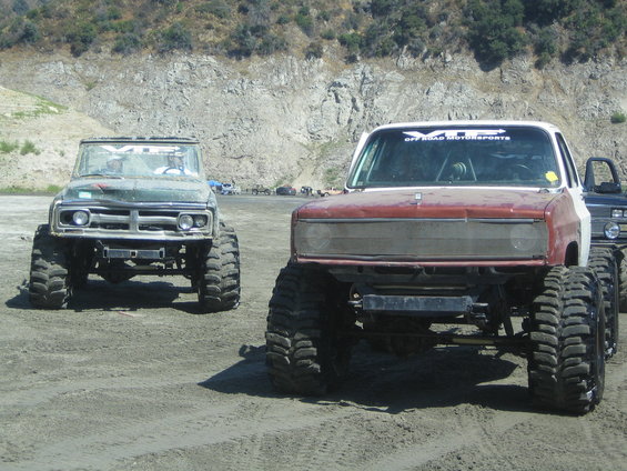 Front view of two lifted off-road trucks with large tires on a sandy surface