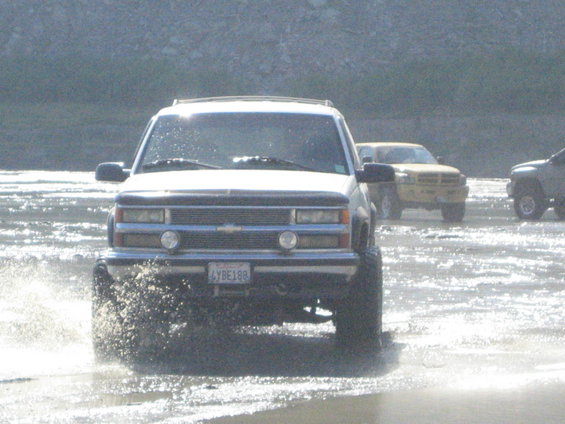 White Chevrolet Tahoe driving through shallow water, kicking up spray. Yellow Dodge Ram pickup and another SUV visible behind it