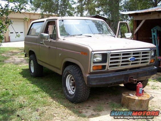Brown 1980 Ford Bronco with white wheels and aftermarket tires parked on grass near a garage