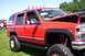 2006 Ford Supercrewzer - This red SUV, likely a Chevrolet Tahoe from the 1990s, features a lifted suspension, large off-road tires, and auxiliary lights on the roof.