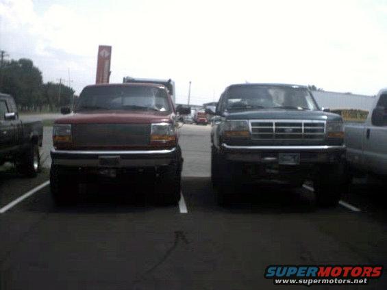 Two lifted pickup trucks, one red and one dark green, parked in a lot. The red truck is on the left