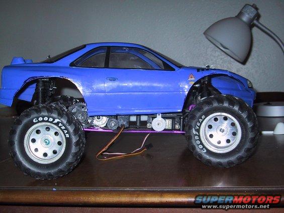 Blue RC monster truck with large tires and exposed gears. A desk lamp is visible in the background