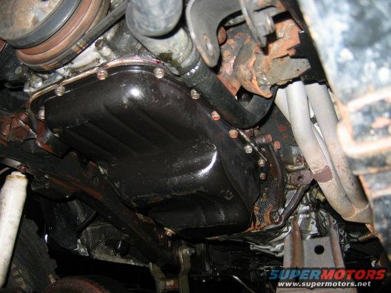 Underside view of an engine oil pan and exhaust manifold with visible rust and hoses