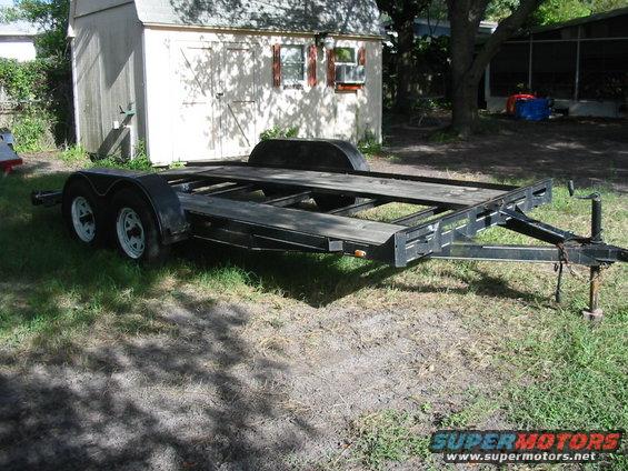 Car hauler trailer with wooden deck and dual axles, parked on grass in front of a shed