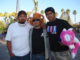 Three guys posing for a photo at an amusement park, one wearing a beer hat and another holding a stuffed animal