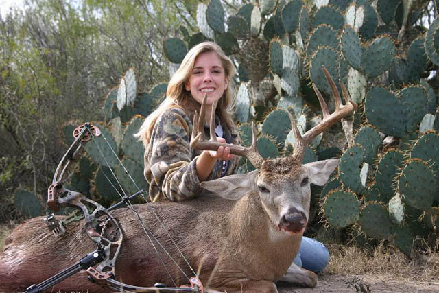 Woman in camouflage sits with a harvested deer and a compound bow in front of prickly pear cactus