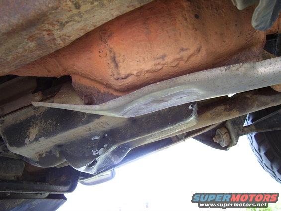 Underside of a vehicle showing rusty frame, exhaust pipe, and suspension components. A metal skid plate is visible