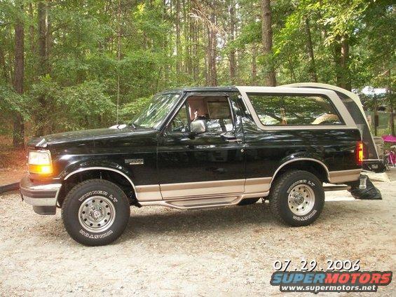 Black Ford Bronco parked on gravel with trees in background. Has tan trim and a removable top