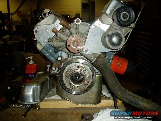 Close-up of a V8 engine block with a water pump, pulley, and oil filter visible. A chrome valve cover sits to the left