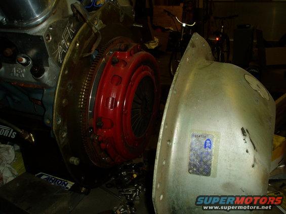Engine clutch assembly with red pressure plate and silver housing, next to a metal shield with a sticker