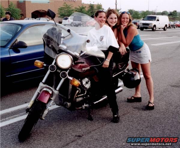 Three young women pose with a motorcycle parked next to a blue car