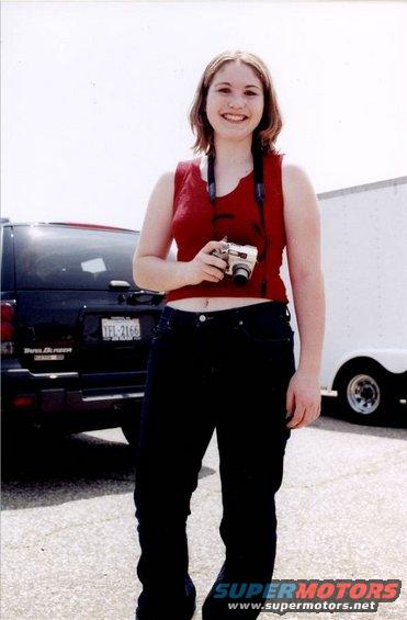 Woman in red top and jeans holds a camera, standing in front of a dark SUV and a white trailer