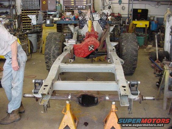 Vehicle frame on jack stands with engine and red transmission assembly. Large tires visible