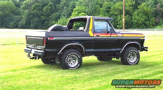 1979 Ford Bronco parked on grass, rear quarter view. Black with yellow, orange, and red stripe