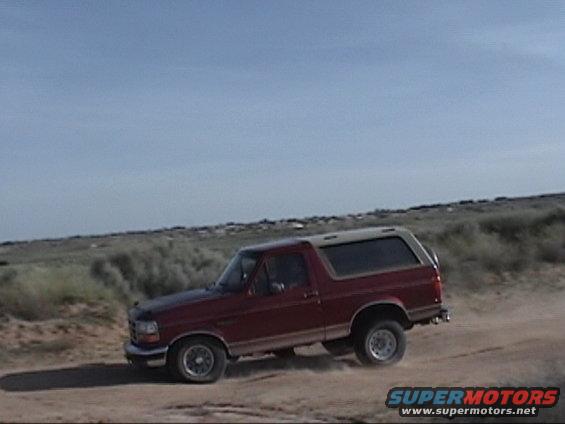 Red SUV driving on a dirt road with a dusty trail behind it. The sky is blue with some clouds