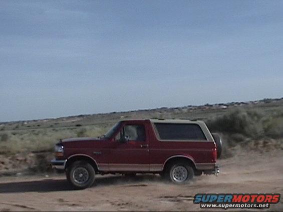 Red 1994 Ford Bronco driving on a dirt road with a spare tire on the back