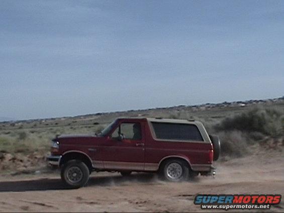 Red Ford Bronco driving on a dirt road, kicking up dust. Spare tire mounted on the rear