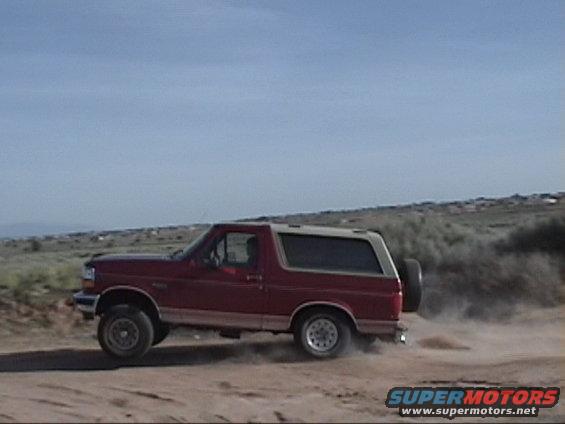 Red Ford Bronco driving on a dirt road, kicking up dust. Spare tire mounted on the back