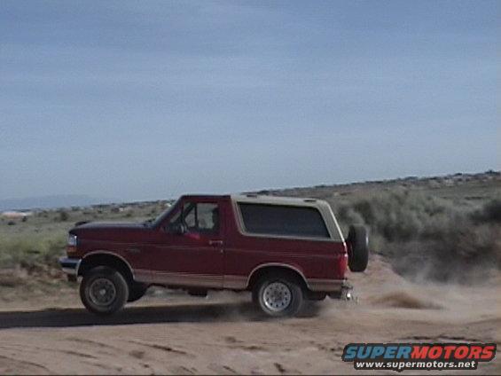 Red Ford Bronco with a tan top, kicking up dust on a dirt road. Spare tire mounted on the rear