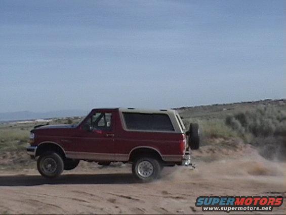 Red 1994 Ford Bronco driving on a dirt road, kicking up dust. Spare tire mounted on the rear